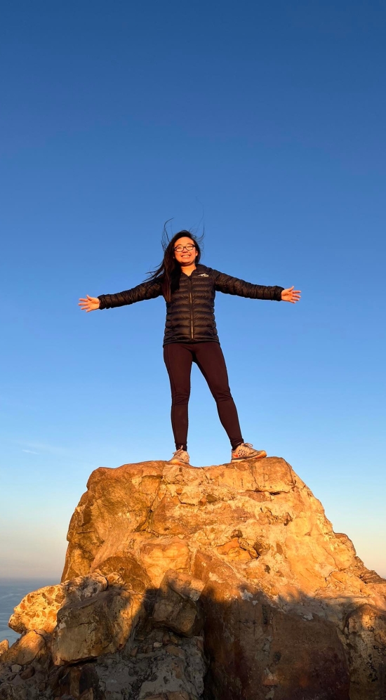 Megan Lim standing on top of a rock posing with two arms wide with the large expanse of blue skies during a sunrise hike up Lion's Head Mountain in South Africa
