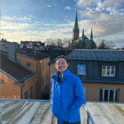 Veronica on a study abroad to Sweden standing with her hands in her purple jacket pocket on a roof that overlooks the Uppsala Cathedral which has green French-inspired spires