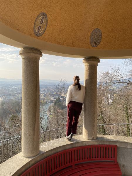 Aquila Simmons with her back to the camera, leaning to her side on a column of a pavilion overlooking the town of Thun with a birds eye view of the town