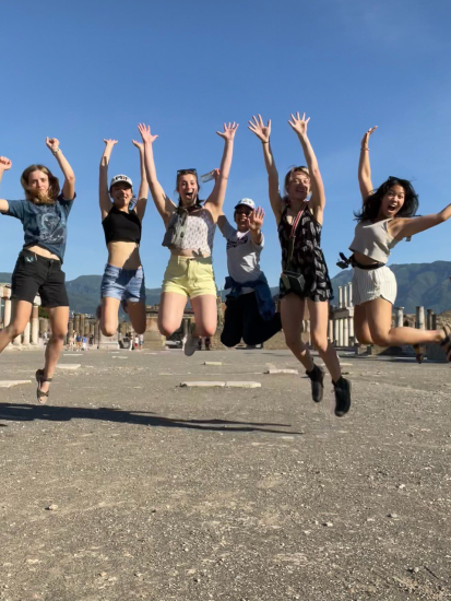 Lainie Beauchemin jumping with her hands up, 2nd to the right with five other MIT students who were in shorts and 2 in caps on a hot sunny day in Pompeii
