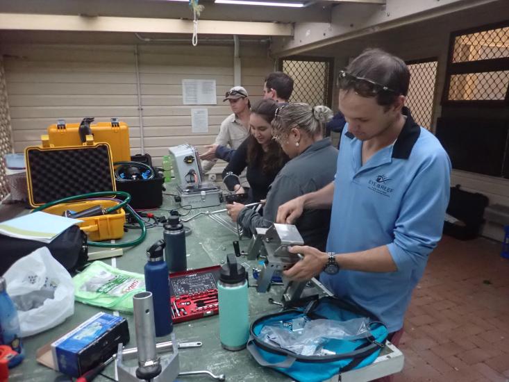 Annemarie Dapoz in a reef scan assembly line with 4 other co-workers handling parts of a reef scan on a table filled with equipment and water bottles