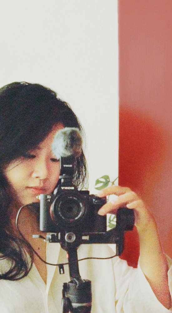 Daisy Zhang holding a camera on a tripod, taking her own photo of herself in a mirror behind a smooth red wall