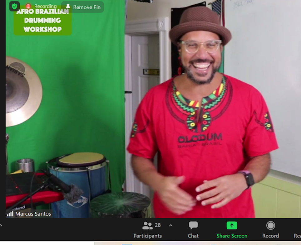 Over Zoom, a Brazillian teacher in red shirt and a drum set in the background teaching Manuel drumming