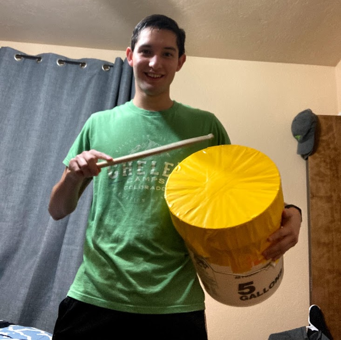 Manuel Morales holding a makeshift drum of a five gallon plastic paint bucket with a yellow plastic wrapped tightly on top of the bucket. He's holding it in one arm and hollding a drum stick in his other hand in his room with a blue curtain drawn behind him.