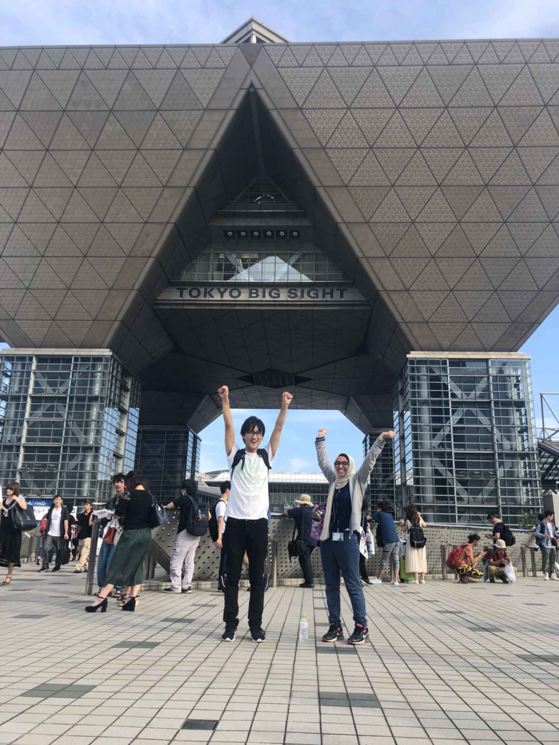 Marwa AlAlawi standing beside her supervisor Ken-san in front Tokyo Big Sight in Odaiba, two inverted pyramids suspended over a plaza.
