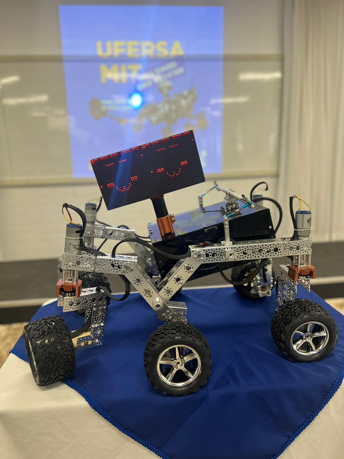 A open source rover on a table with 6 wheels and a lined metal body with a rectangular screen that has two smiley faces