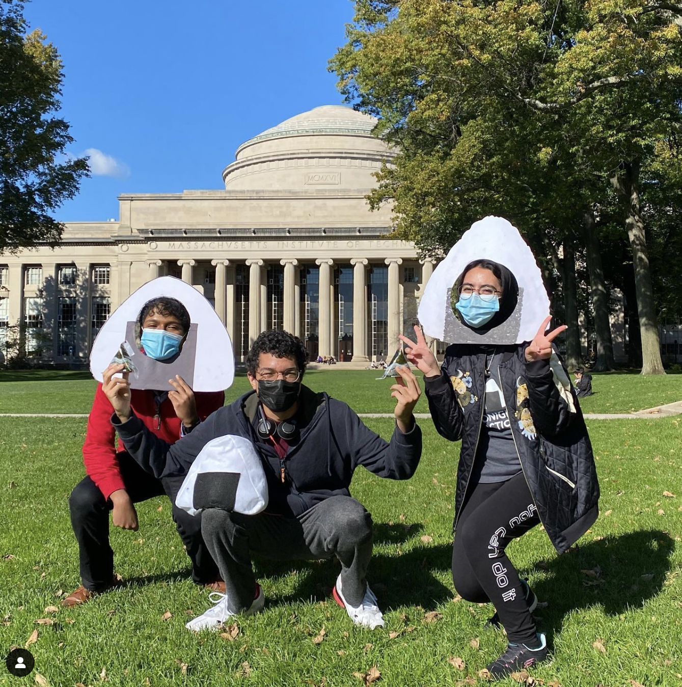 Marwa AlAlawi wearing a onigiri photobooth prop holding two peace signs with both hands crouching down halfway to the right with two other MIT students who's also crouching down on the grass, one wearing a onigiri photobooth prop and another with glasses holding an onigiri rice ball in one hand and rested on his thigh, an onigiri pillow in front of Killian Court at MIT