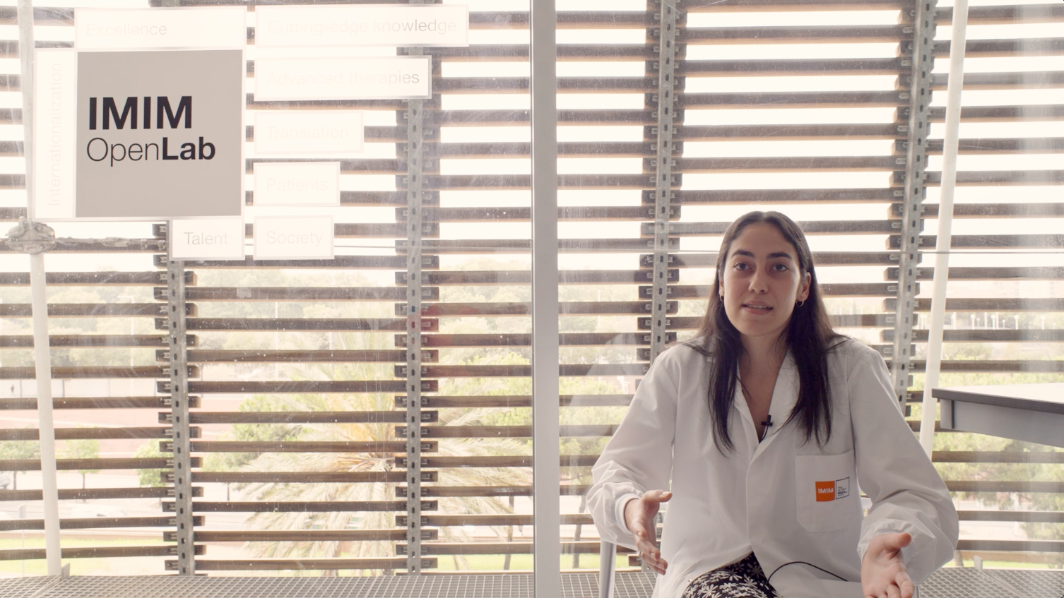 Elizabeth Carbonell '23 sitting down to the right of the screen in a lab coat doing an interview at the IMIM Open Lab office 