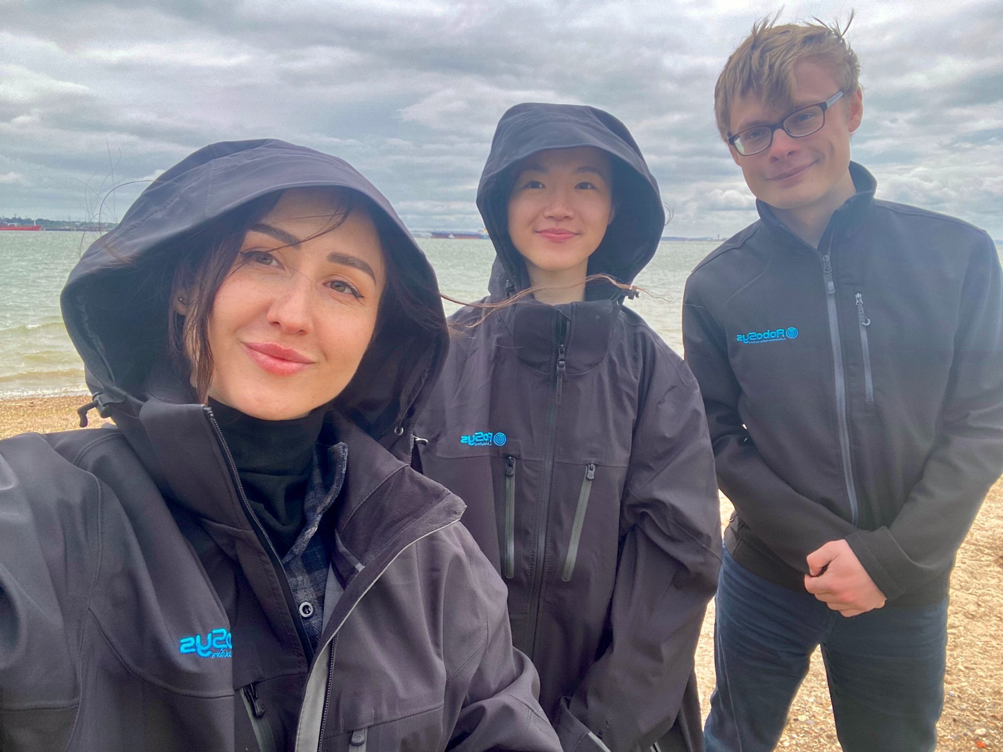 In a selfie taken by Amy Shi's colleague, Amy stands between two of them on a beach on a cloudy day wearing the company's hooded black jacket