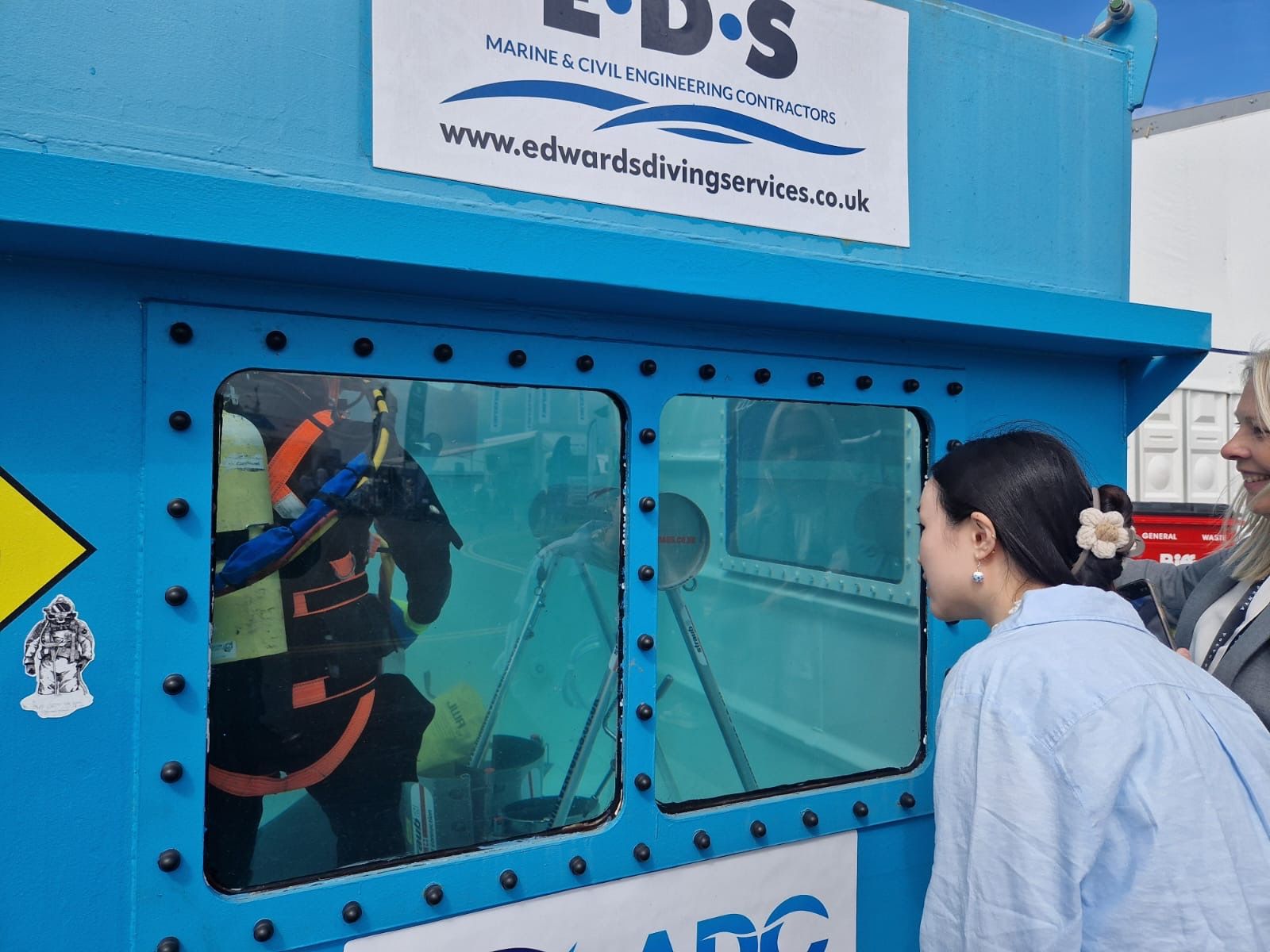 Amy Shi looking into a blue tank with someone in it wearing a wetsuit and oxygen tank under water