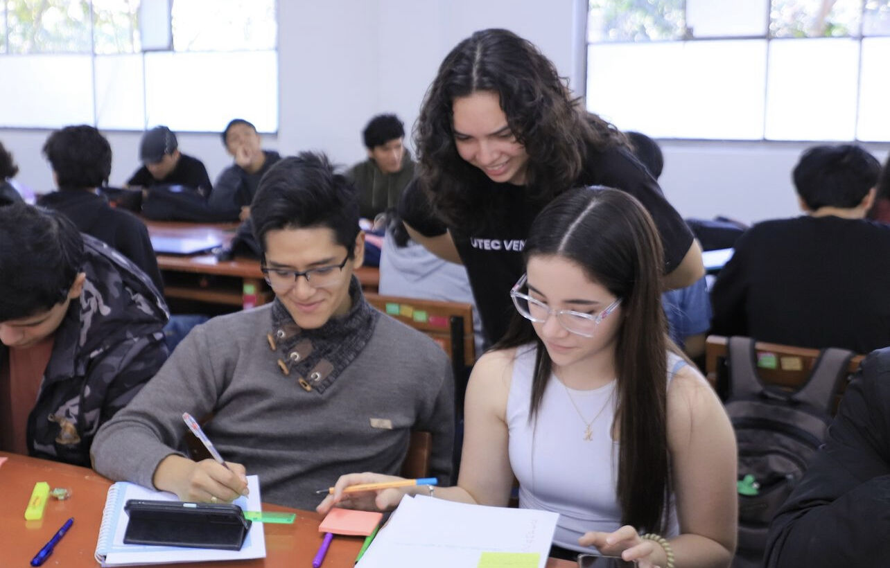 Ana Tejeda standing over two students in Peru while they look at their books at a brown table in a bright lit class with other students