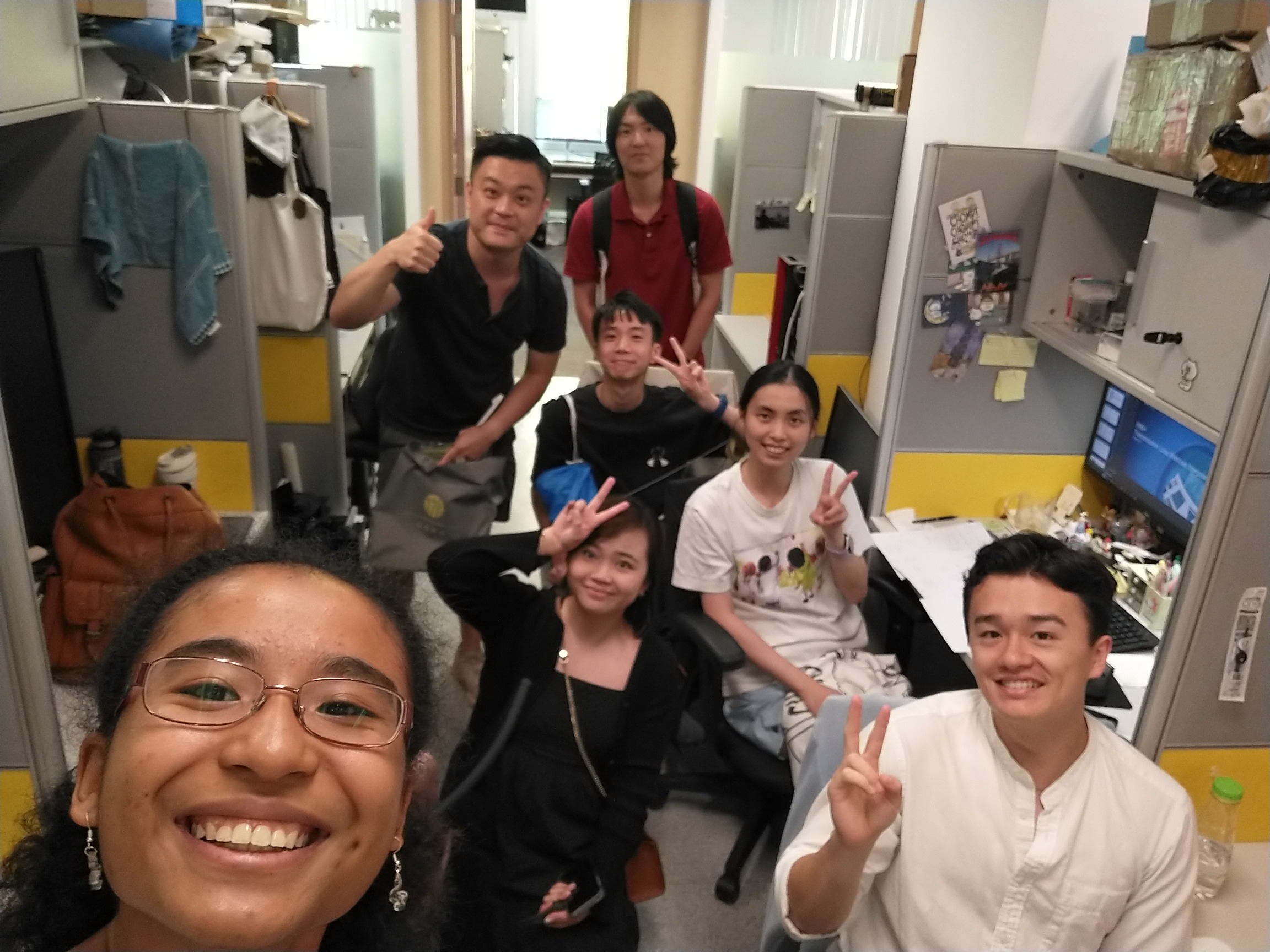 Jolie Duquene taking a selfie with coworkers in Hong Kong University. Three seated on chairs holding peace sign and others standing around narrow office flanked with cubicles on both side