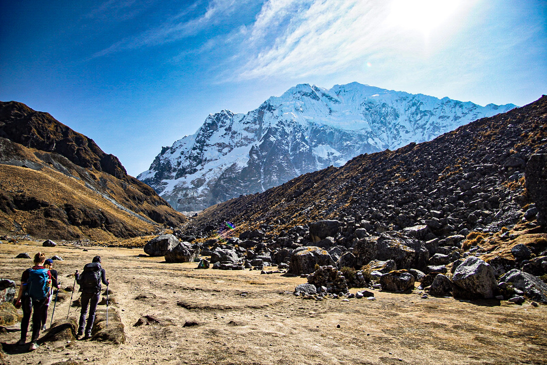 The image shows a mountainous landscape with a clear sky. In the foreground, two hikers with backpacks and trekking poles walk along a rocky path. The ground is a mix of dry grass and scattered rocks. Behind them rise steep, rocky hills, partially covered with golden-brown grass. Prominently in the background is a massive snow-covered 