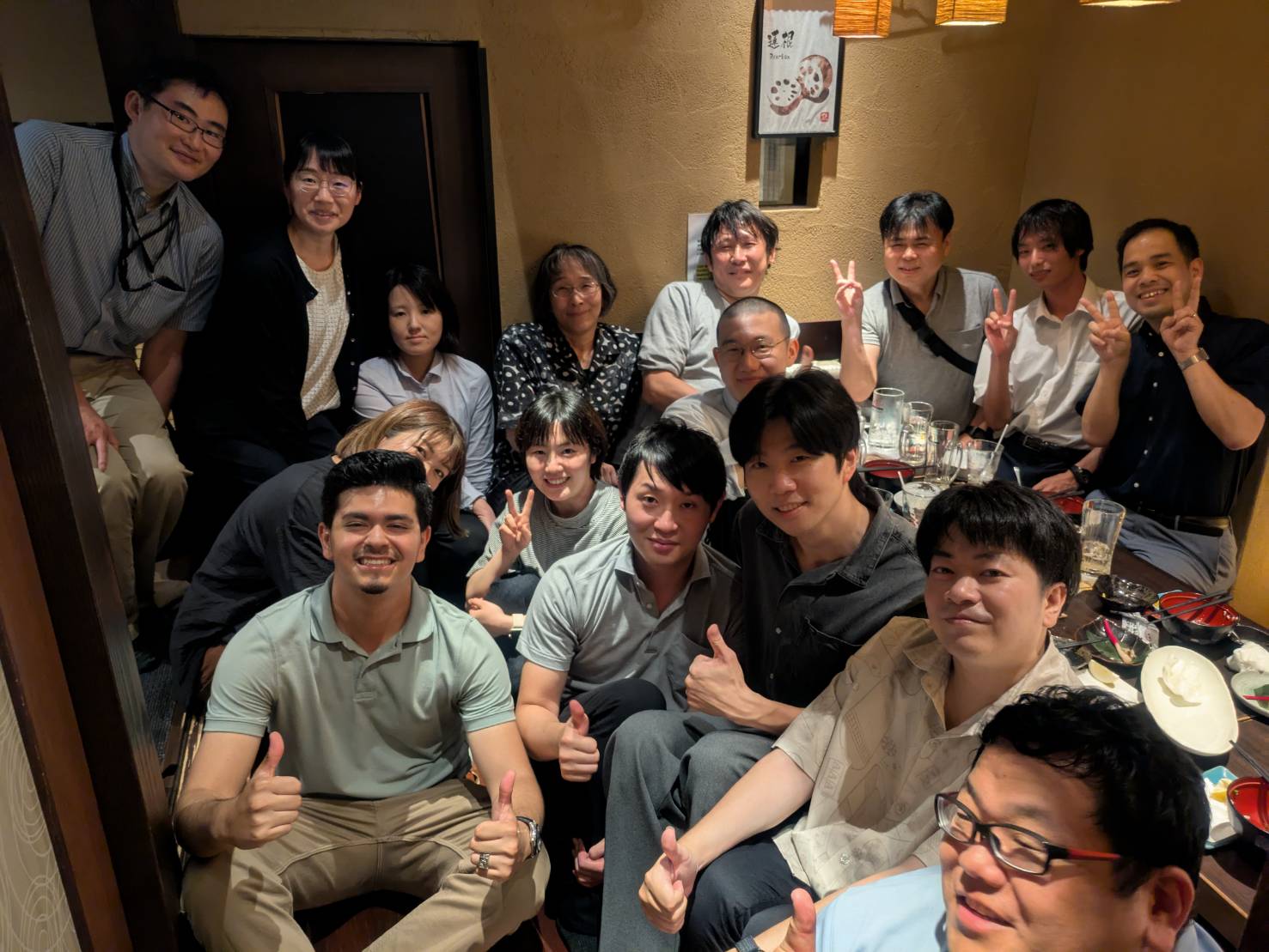 Daniel and 15 others huddled in a room with some holding thumbs up, peace signs in a room