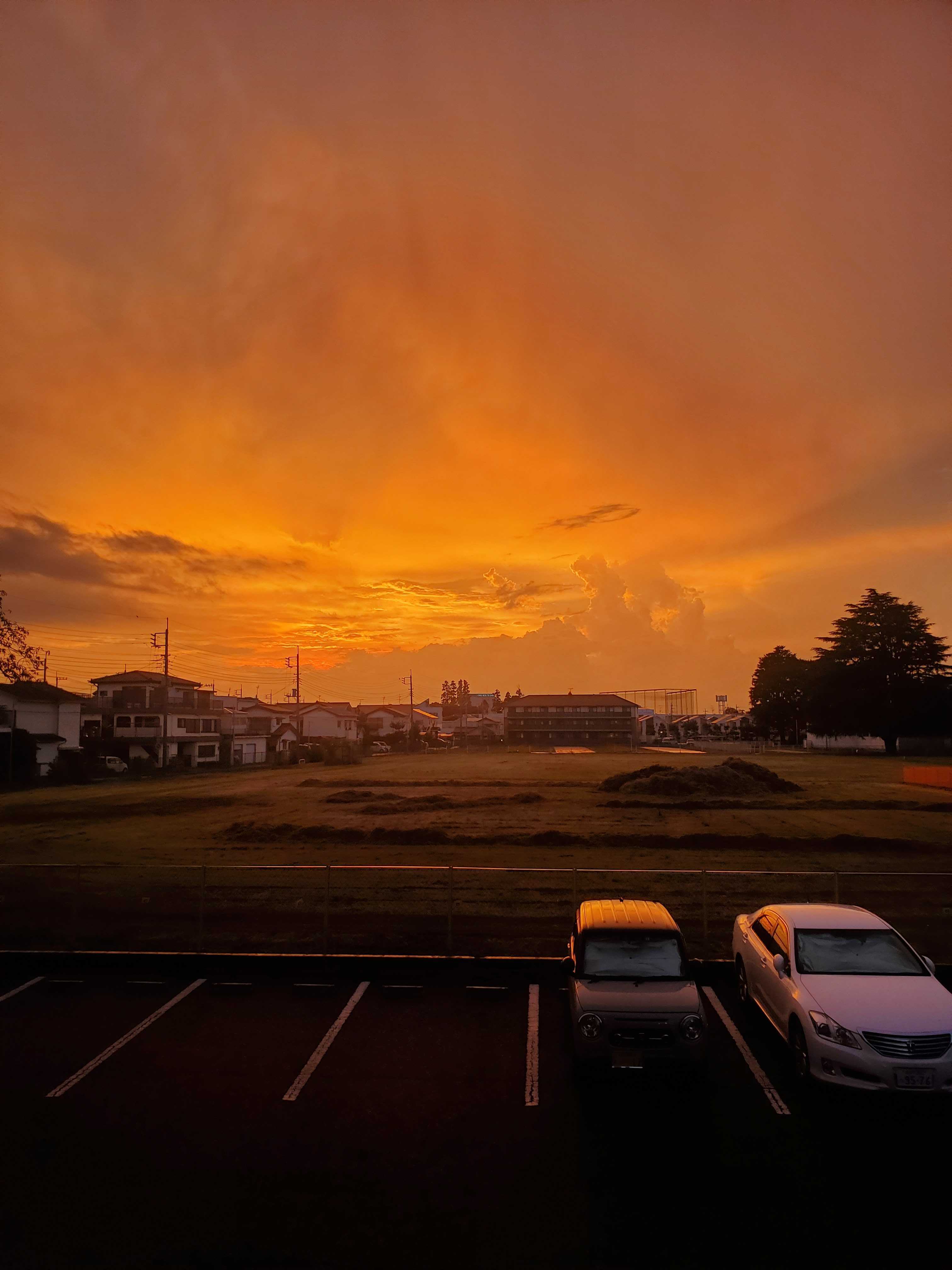 Sunset over a suburban landscape with orange sky and clouds, cars in a parking lot, and residential buildings in the background in Hitachinaka, Japan