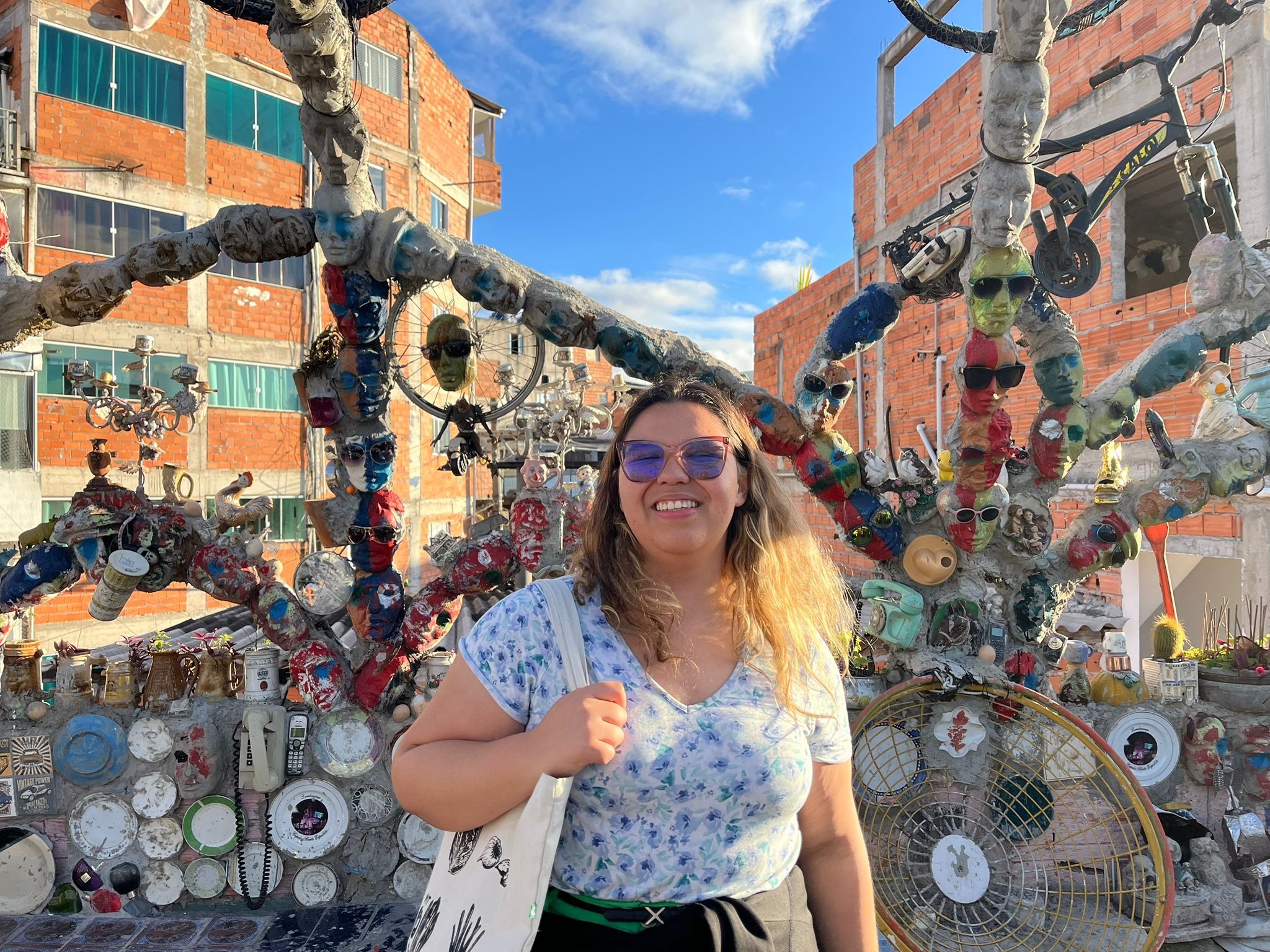 Hana Sousa wearing sunglasses holding onto the strap of her white tote bag in front a structured art piece on a bridge