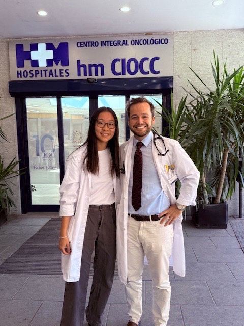 Yolanda Xie, MIT course 6, standing next to supervisor, and both wearing white lab coats in front of the entrance to HM Hospitales in Madrid, Spain