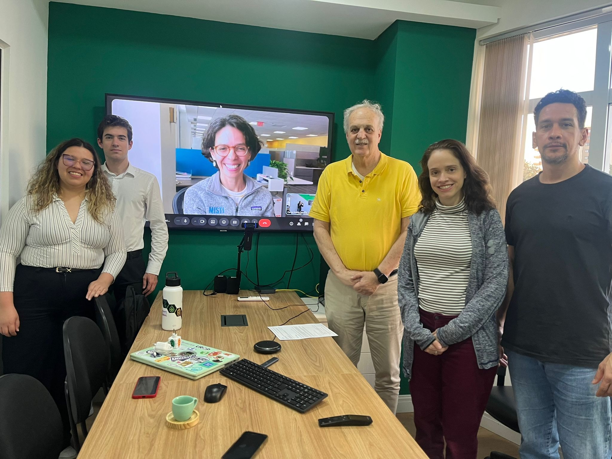 Hana Sousa standing to the far left in the corner with a another colleague to the left of a rectangle wooden table in a conference room with Rosabelli on the screen in the middle and 3 other colleagues to the right of the table
