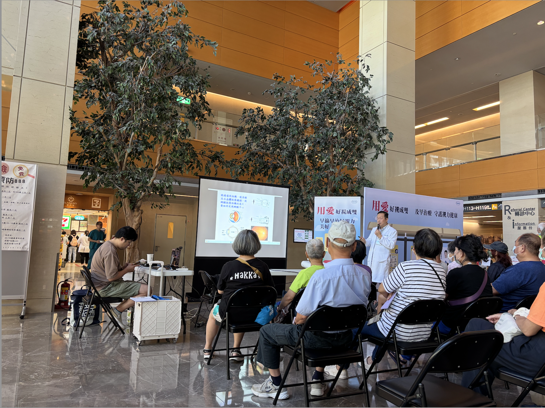 Hospital lobby with two towering trees from the first floor reaching up to the second with locals seated on foldable black chairs in front of a presentation by a person in a doctor's white coat holding a mic in FarEastern Hospital, Taiwan, taken by Kathy Hu
