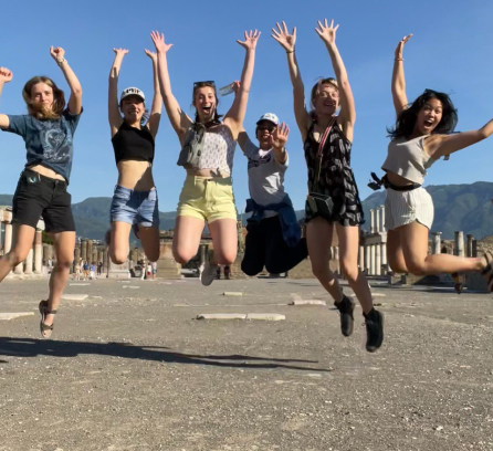 Lainie Beauchemin jumping with her hands up, 2nd to the right with five other MIT students who were in shorts and 2 in caps on a hot sunny day in Pompeii
