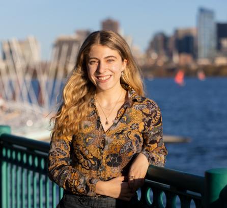 Aviva Intveld, smiling and leaning on a railing overlooking the Charles River with the Boston skyline in the background