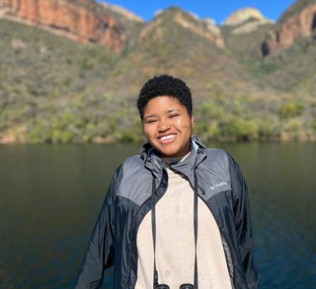 Elissa Gibson standing in front of a lake with mountains in the background. The sky is blue and cloudless. She is smiling and wearing binoculars around her neck.