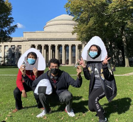 Marwa AlAlawi wearing a onigiri photobooth prop holding two peace signs with both hands crouching down halfway to the right with two other MIT students who's also crouching down on the grass, one wearing a onigiri photobooth prop and another with glasses holding an onigiri rice ball in one hand and rested on his thigh, an onigiri pillow in front of Killian Court at MIT