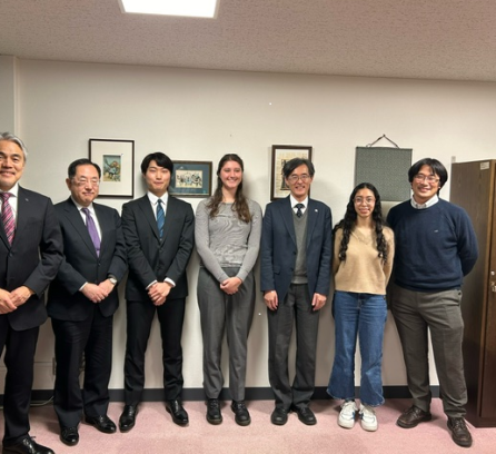 Riley Sage Moeykens in a long grey sweater and Carmen Avila in a brown sweater standing amongst Tokyo Tech EVP Imura and Tokyo Tech-MIT Student Exchange Program Committee members clad in suits and Prof. Takuya Harada in a sweater and collared shirt