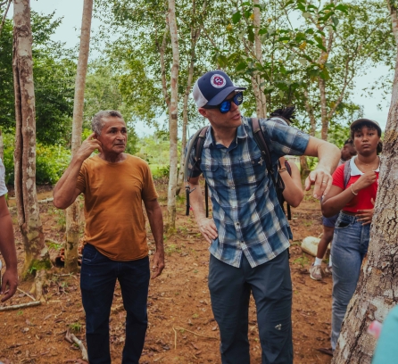 Professor Brad Olsen in a baseball cap looking at a tree, students from MIT and MUSA, surrounding him in the Amazon