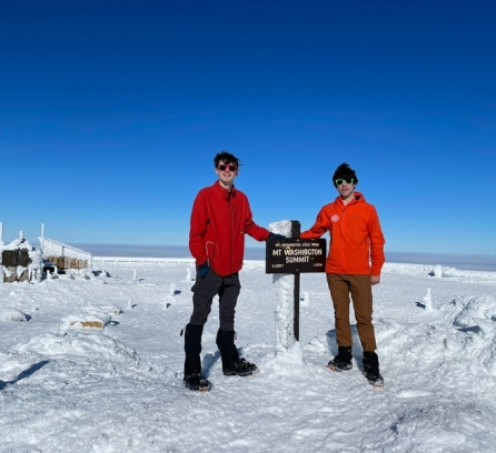 Nathanael Jenkins and Timur Uyumaz pose with the summit marker on Mt. Washington on a sunny day. They wear cold-weather gear and there is snow on the ground.
