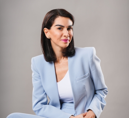 Maye Elqasem headshot in a light blue outfit seated with a grey background 
