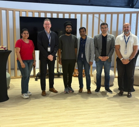 Smruthi Kilari, Andrew Murphy, Amitoj Singh, Satish Kumar, Rajeet Sampat, and Wesam Omar pose standing indoors with a blank TV screen behind them.