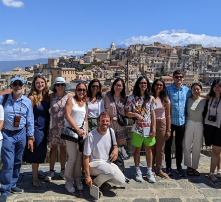 16 people pose on a plaza overlooking an Italian village with mountains in background