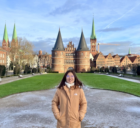 Crystal Liang, wearing a winter jacket, stands in front of a German castle with round towers and high, pointed spires