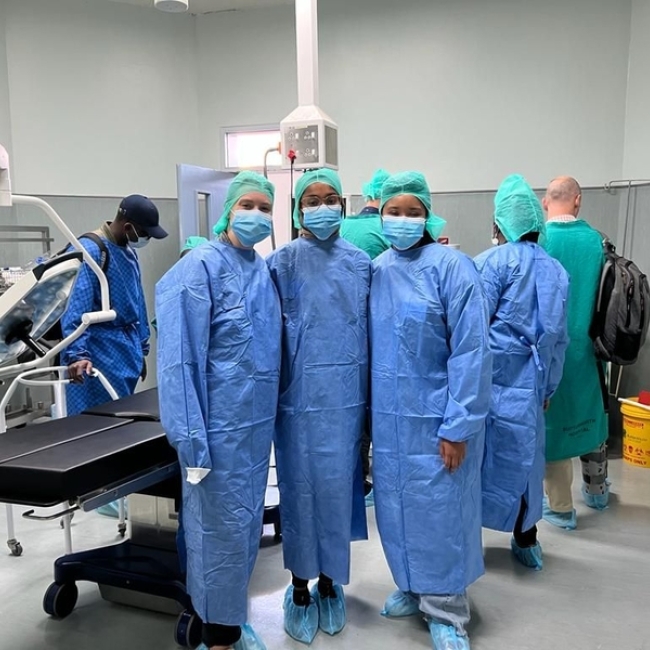 Three young adults are photographed in full scrubs — blue head covers, blue face masks, blue gowns, and blue shoe coverings — in a surgical room of a hospital. Elissa Gibson in on the right. Four other people can be seen in the background. The room is white, grey, and sterile looking. An empty black surgery table is behind the group.