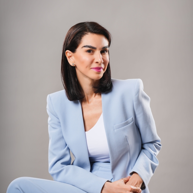 Maye Elqasem headshot in a light blue outfit seated with a grey background 