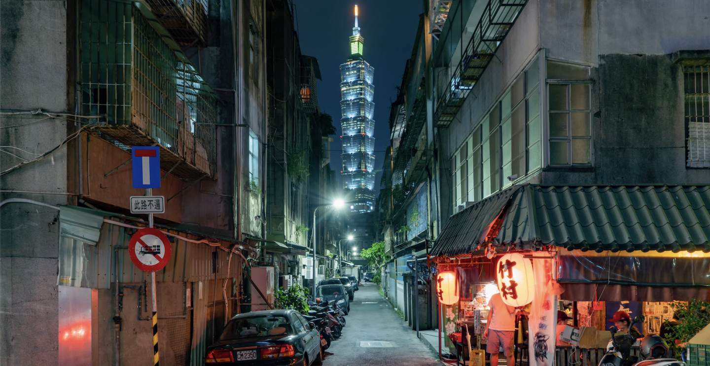 Alley way in the night with cars lined to the left and a shop to the right corner with red round Chinese lanterns and the Taipei Tower seen in a distance