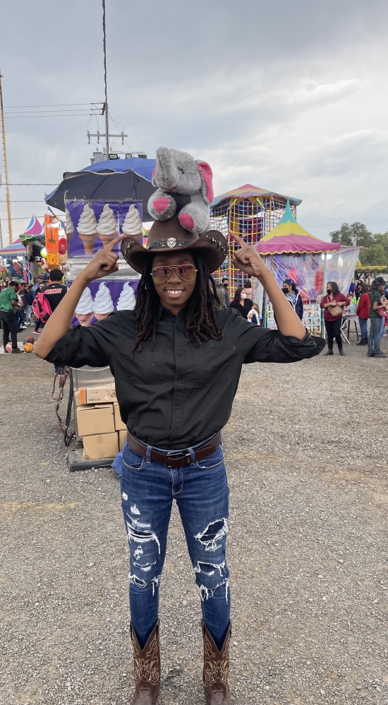 Aniesha Dyce pointing her fingers to a cowboy hat she's wearing in a fair in Mexico. Colourful tents in the fair in the background