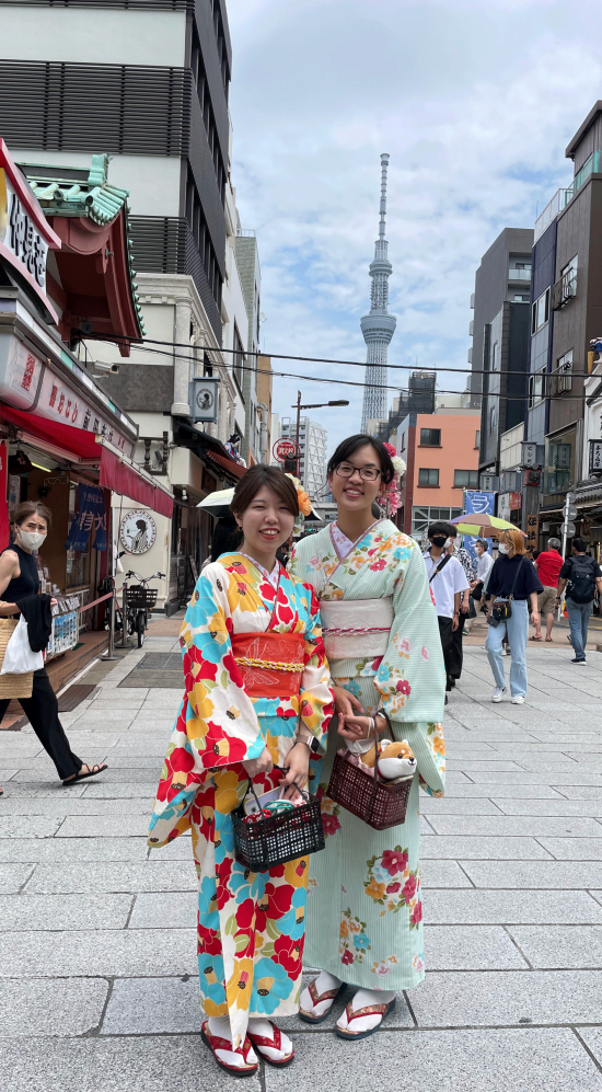 Demi Fang donning a kimono with her friend with a row of shops in the background and the Tokyo Tower in view in the background with an overcast