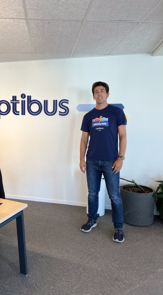 Jonathan Berger standing to the right of a optibus logo decal against a white wall with a brown wooden-table top to the left with a monitor and chair to the left of the picture