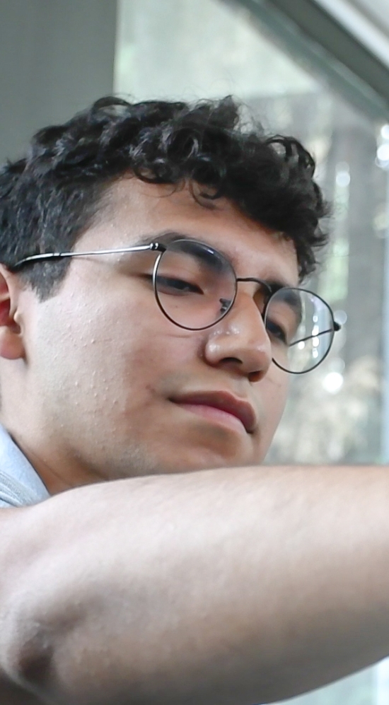 Close up of Maximiliano Ramirez using one hand to hold a glass stirring rod that's in a glass beaker. A window with blinds and dark grey walls in the background