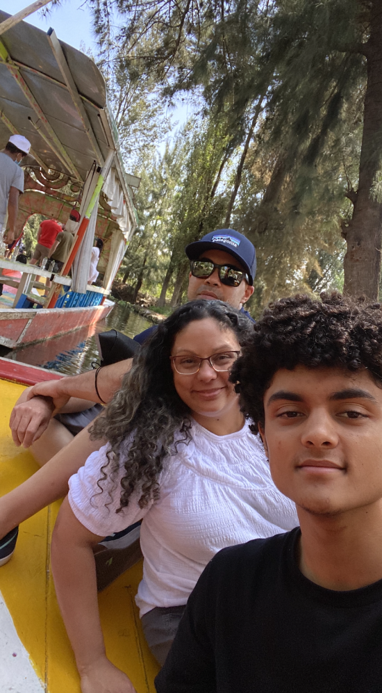 Diego Barros taking a selfie with his parents at the back on a boat and a background of trees and two men standing in a boat behind them