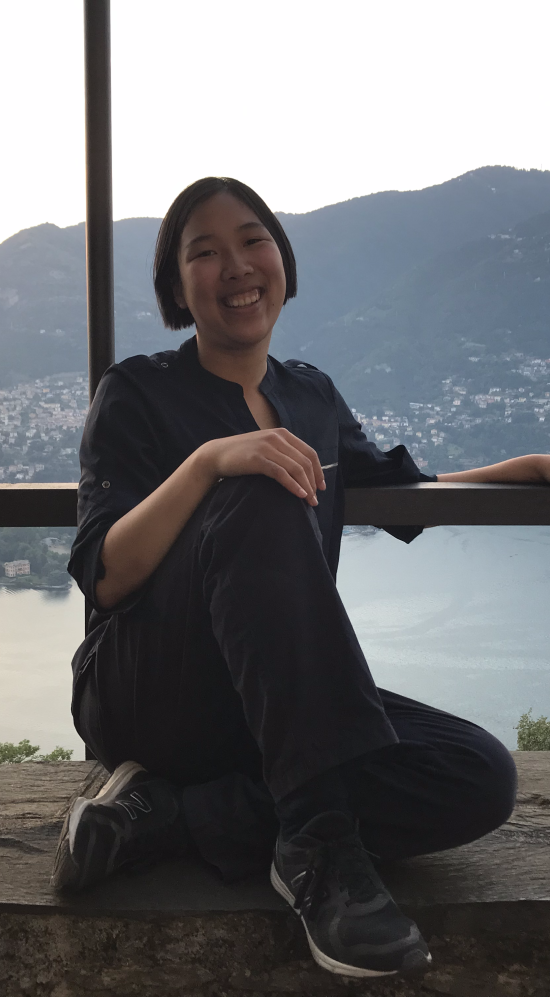 Melbourne Tang sitting down leaning on a single rail with Lake Como, a birds-eye view of the city of Como and mountain ranges