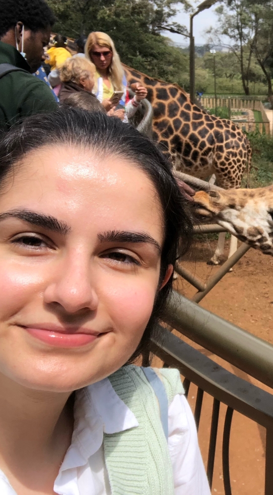 A selfie of Negin Amouei and a giraffe behind a railing to the right, being fed by other tourist