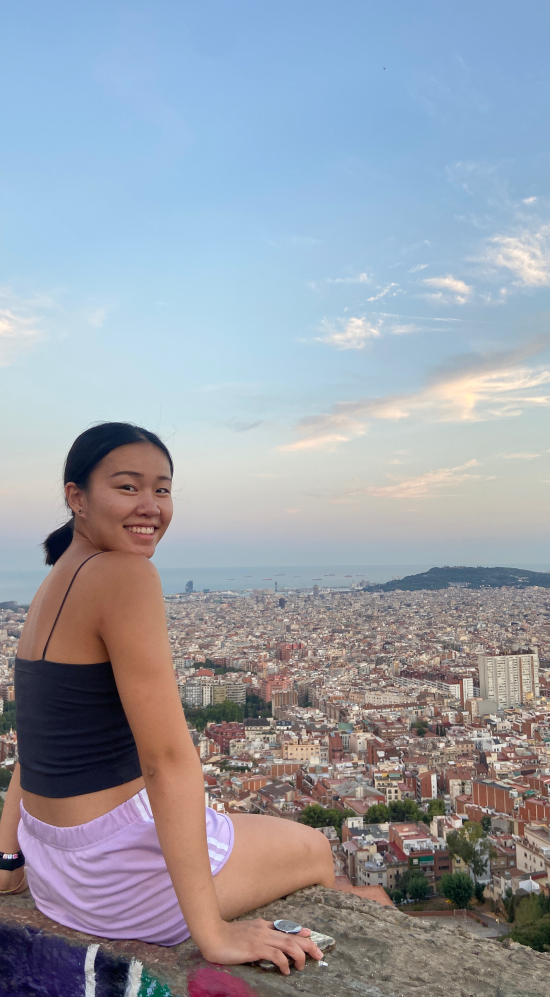 Sally Zhao sitting on the hilltop ruins of the Bunkers del Carmel while looking back to smile at the camera with a bird's-eye view of Barcelona, Catalonia in the background