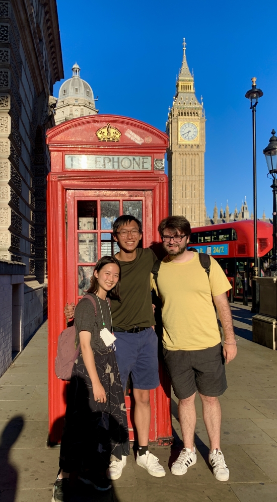 Tiffany Louie standing far left with two other MIT students in front of a red phone booth in London with Big Ben, a red bus and street lamps along the sidewalk in the background