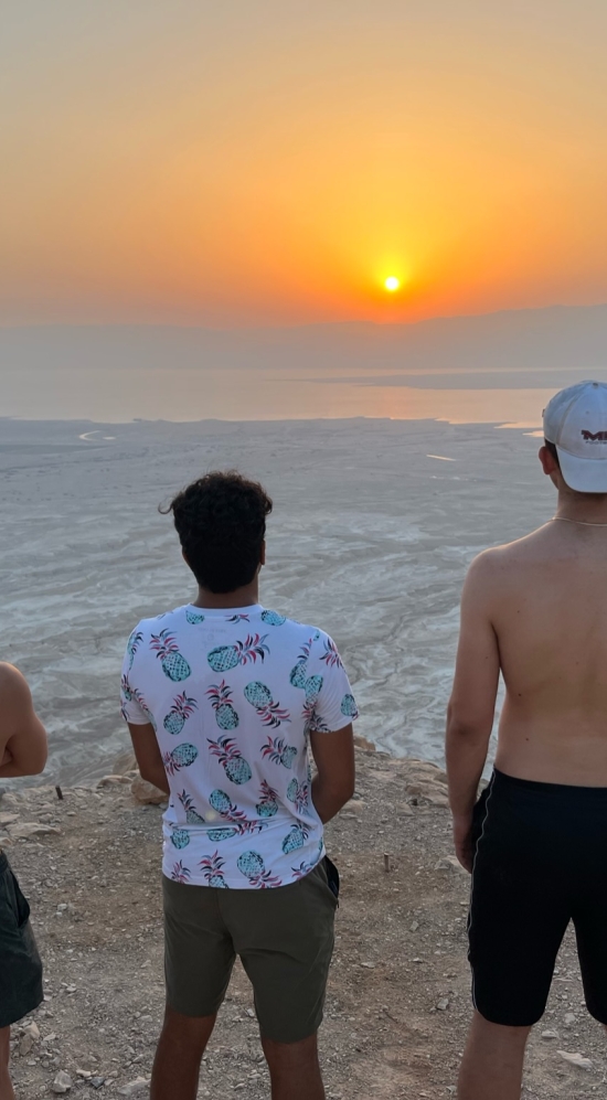 Yajvan Ravan standing in the middle of two other MIT students on top of Masada fortress in Israel, overlooking the Dead Sea and looking out at the sunrise in the horizon