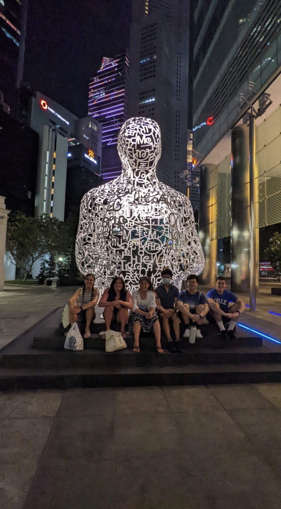Joseph Cahaly with 5 other MIT students sitting in front of a lit up white art work of a person surrounded by city lights and tall sky scrappers in Singapore at night 
