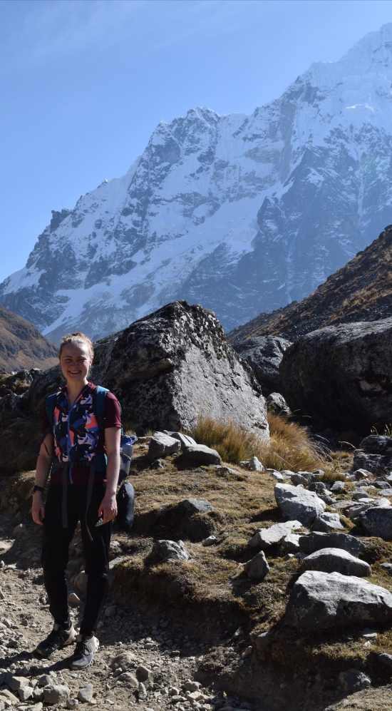 Kira Buttrey hiking the Salkantay Trek with some rock formations along the path and a snow-capped mountainous region in the background