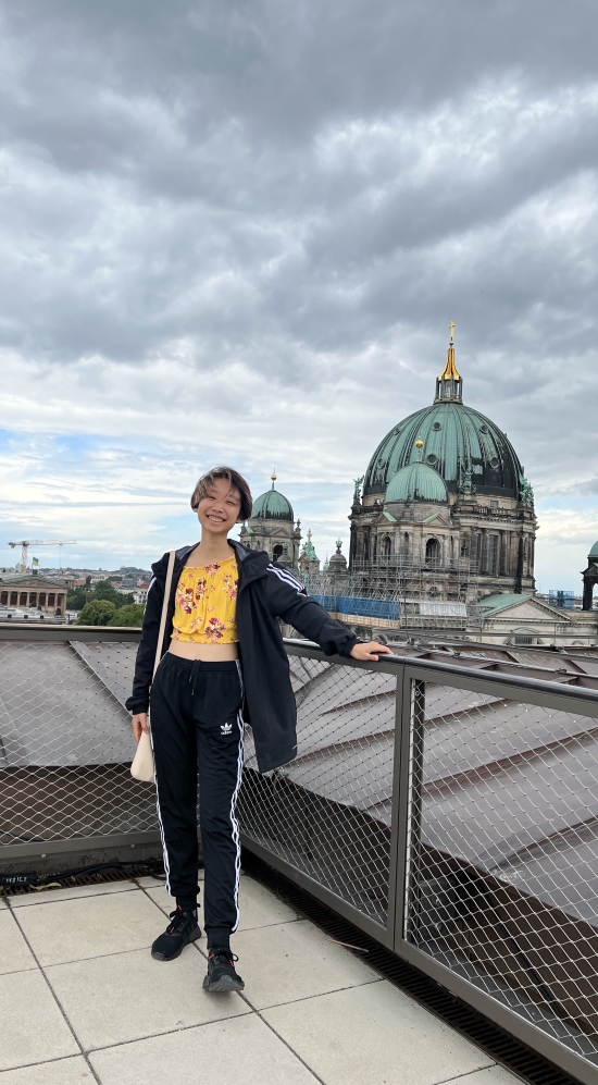 Tian Lin having one hand on a railing with the Berlin Cathedral, a green domed building with a gold spire at the top in the background